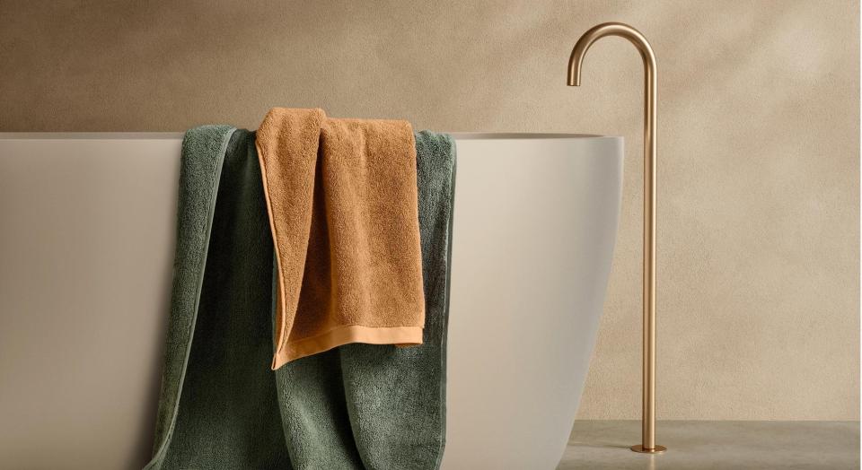 sheridan bath towels in green and yellow draped over white marble bath tub, with gold top, marble floor and beige painted wall in background