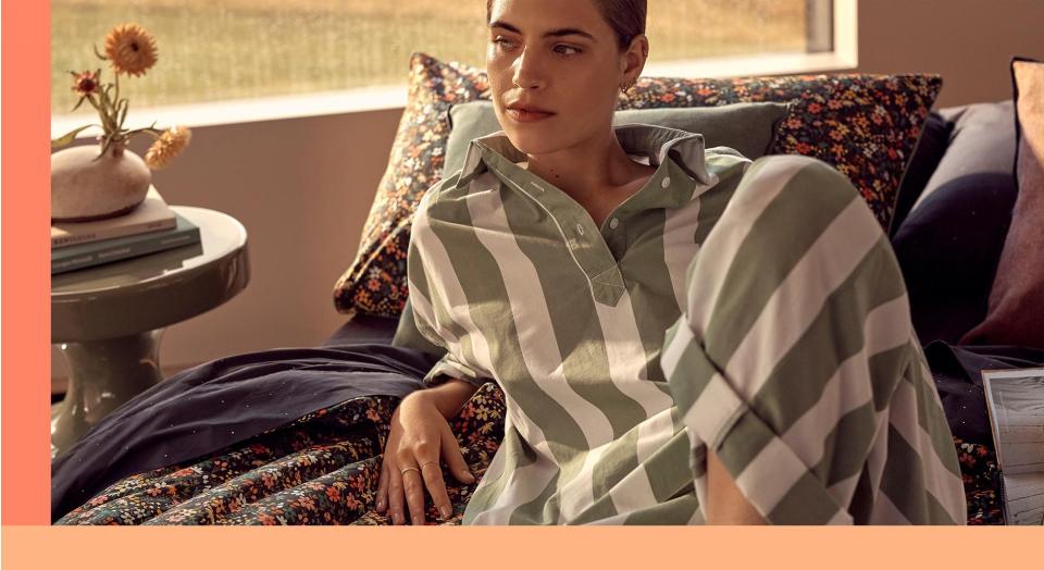70s inspired bedroom decor, where white woman in green and white striped loungewear sits atop of bed covered in ditsy floral print