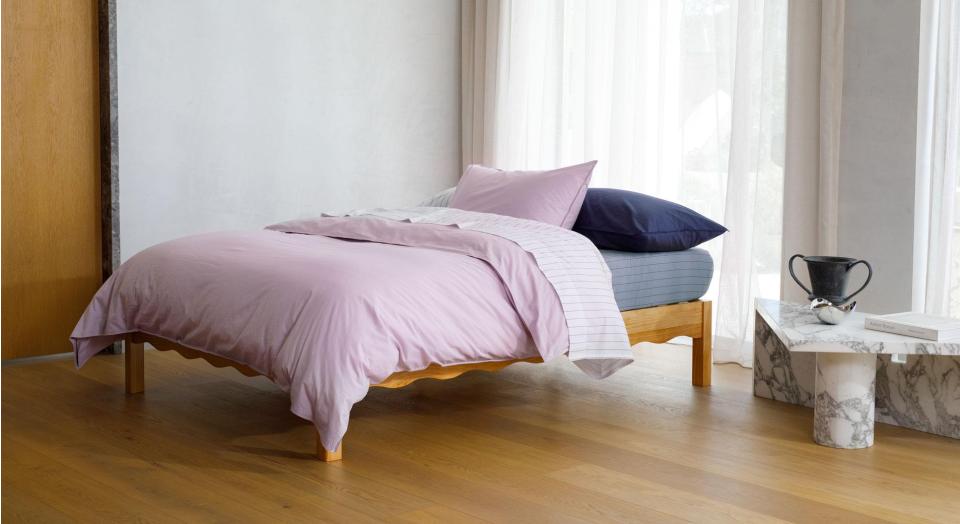 A bed sitting in a modern room with white walls, timber floors and a large window with sheer fabric curtains. The bed is dressed in Bayley Washed Percale bedding in dusty mauve, white, steel blue and deep sea