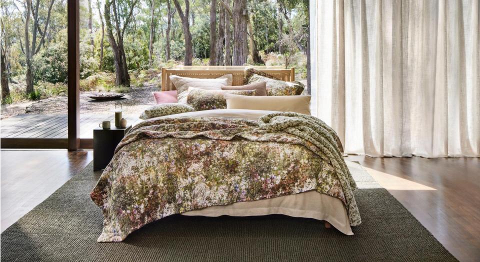 A bed sits in front of a large window with a green rug underneath. Outside is a tree-lined Australian landscape.