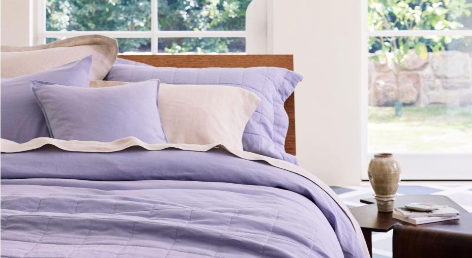 A bed made with violet-coloured bed linen. In the background is a white wall with tall, arched windows and a garden outside. Beside the bed is a timber side table with a small ceramic vase and assorted trinkets on top.