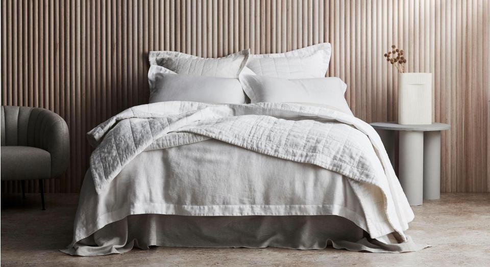 A bed sits in front of a timber wall. On either side of the bed is a fabric armchair and a grey side table with a vase of dried flowers. The bed is dressed in white Abbotson Linen bedding.