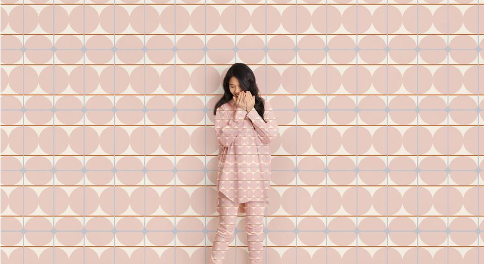 A woman stands, giggling and covering her face in front of a geometric print background. Her shirt and pants are in the same matching pattern.