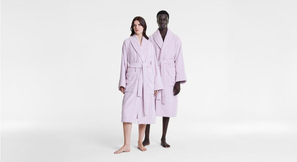 A man and woman stand side by side in front of a blank white set. Both are modelling Aven Stripe bathrobes in lilac.