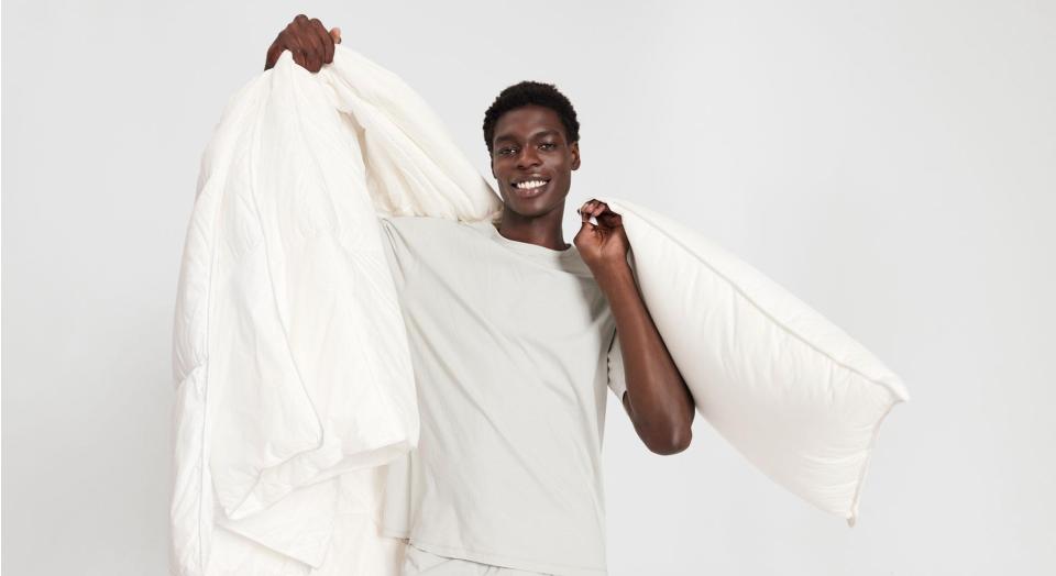 A young man in front of a grey backdrop smiling and holding up a pillow in one hand and a quilt in the other.