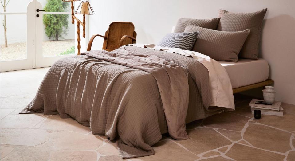 A bedroom with natural stone floors. The bed sits next to a wooden lamp and armchair and is covered in pale pink and soft brown bedding.