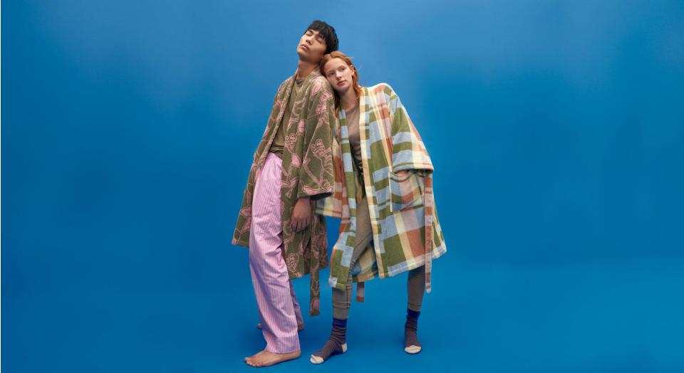 Young man and woman stand in front of a blue backdrop, both wearing stylish bathrobes. 