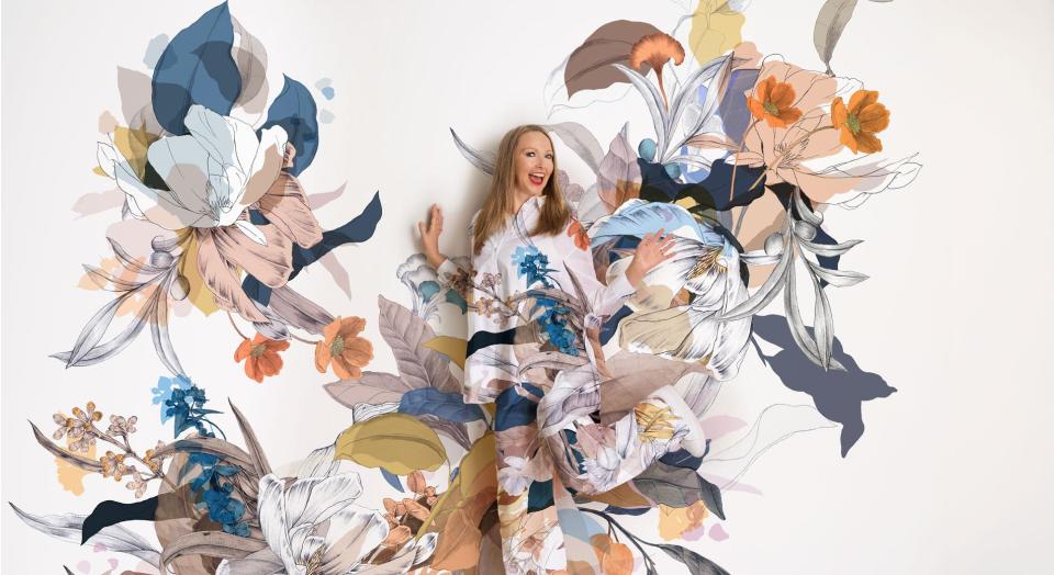 A woman stands, smiling in front of a colourful floral print background. Her shirt and pants are in the same matching pattern.