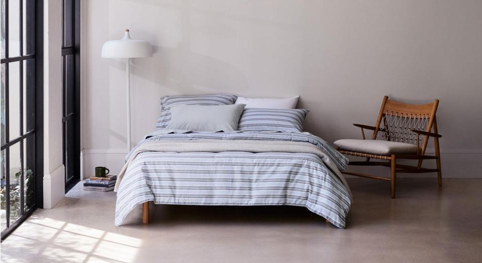 A bed dressed in blue striped flannelette bedding sits in a modern bedroom with concrete floors and French windows. Next to the bed is a wooden armchair, a white lamp and a small stack of books with a mug sitting on top.