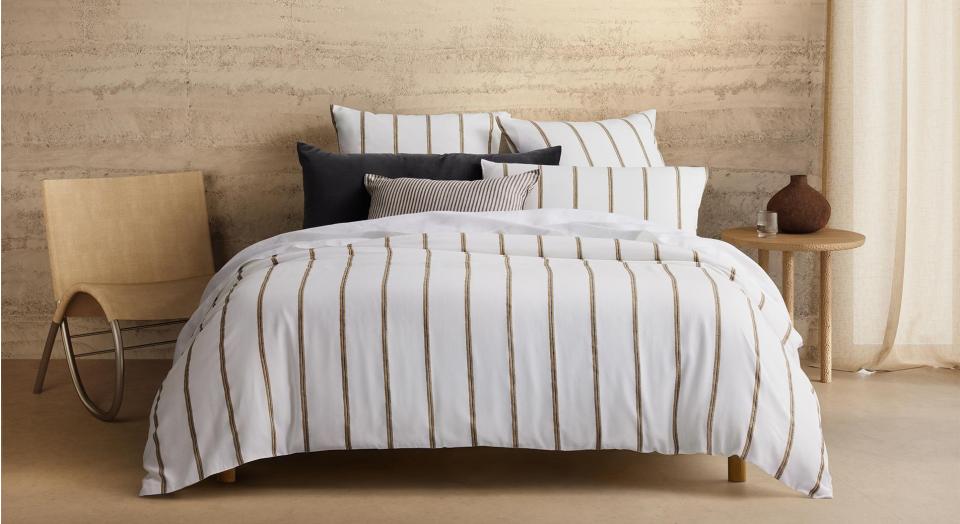 A white bed with brown stripes next to a wicker chair and timber bedside table in a room with beige concrete walls and flooring