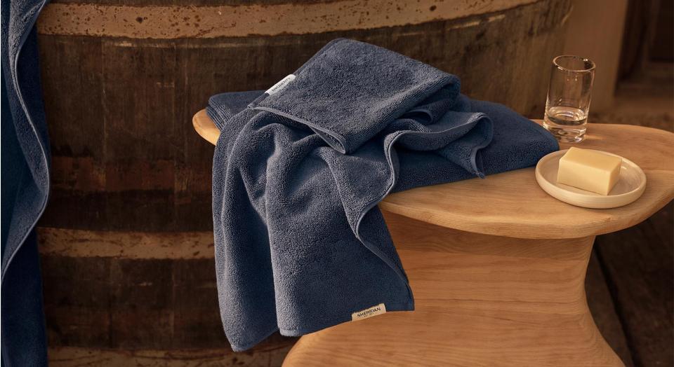 Navy towels draped over a timber side table, next to a glass of water and a bar of soap. A rustic timber bathtub in the background.