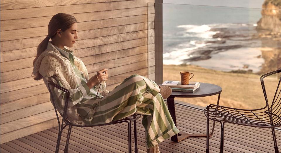 woman in striped green and white loungewear set sits on a balcony outside, with wooden wall and wooden floor, and glass railing. she has a view of grassy knoll, leading down to rocky cliff front and ocean.
