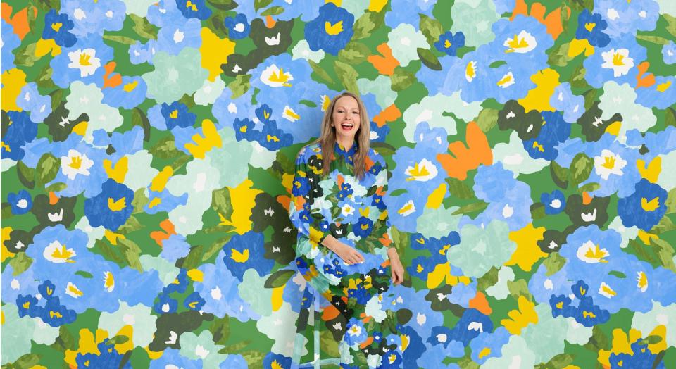 A woman sits, smiling in front of a vivid floral print background. Her shirt and pants are in the same matching pattern.