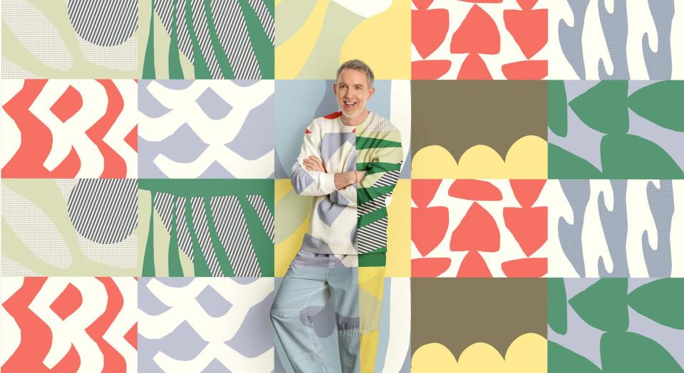 A man stands, smiling in front of a colourful geometric print background. His shirt and pants are in the same matching pattern.