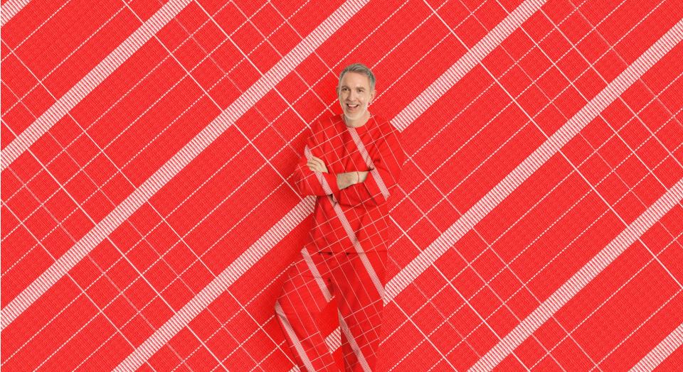 A man stands, smiling with his arms crossed in front of a red and white check print background. His shirt and pants are in the same matching pattern.