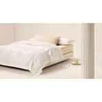 A bed sitting in an empty white room. The bed is dressed in Millennia bedding. On the floor next to the bed is a slice of cake on a plate.