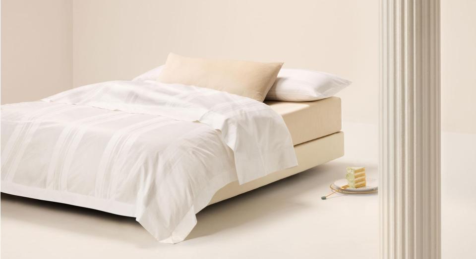 A bed sitting in an empty white room. The bed is dressed in Millennia bedding. On the floor next to the bed is a slice of cake on a plate.