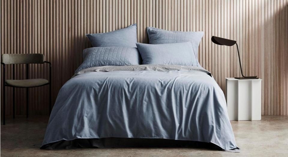 A bed sits in a modern bedroom with timber walls. On either side of the bed is a chair and a white side table with a black metal lamp. The bed is dressed in Reilly chambray bedding.