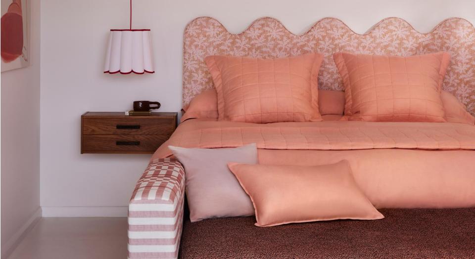 Bed with scalloped fabric headboard and coral pink sheets sits in front of a white wall. There is a bench at the foot of the bed with two pink pillows sitting on top, and a white lamp shade hanging from the ceiling.