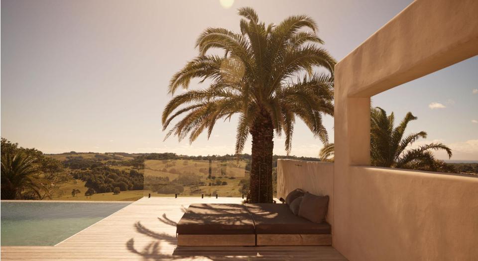 A balcony looking out to grassy hills. In the foreground is a pool and a daybed underneath a palm tree