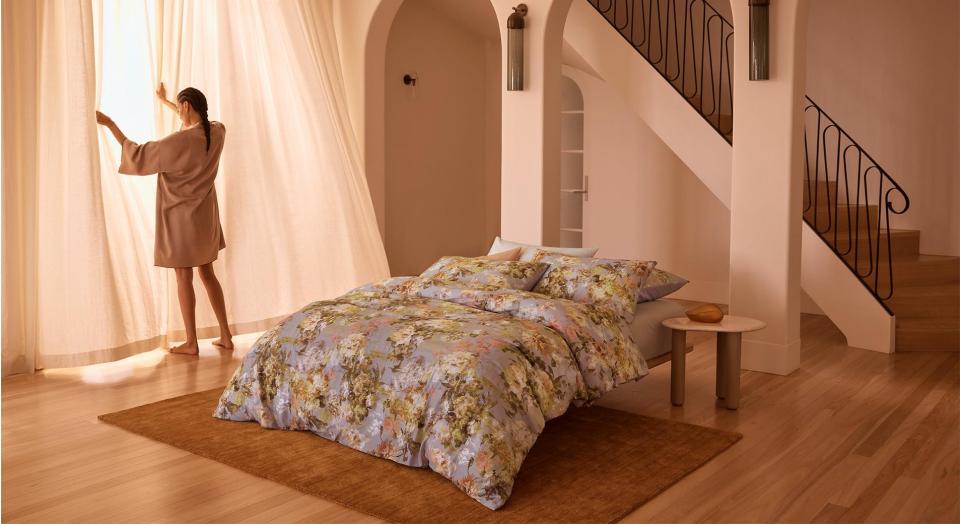 A woman in front of a tall window opening flowing, white curtains. Next to her is a bed with a floral quilt cover.