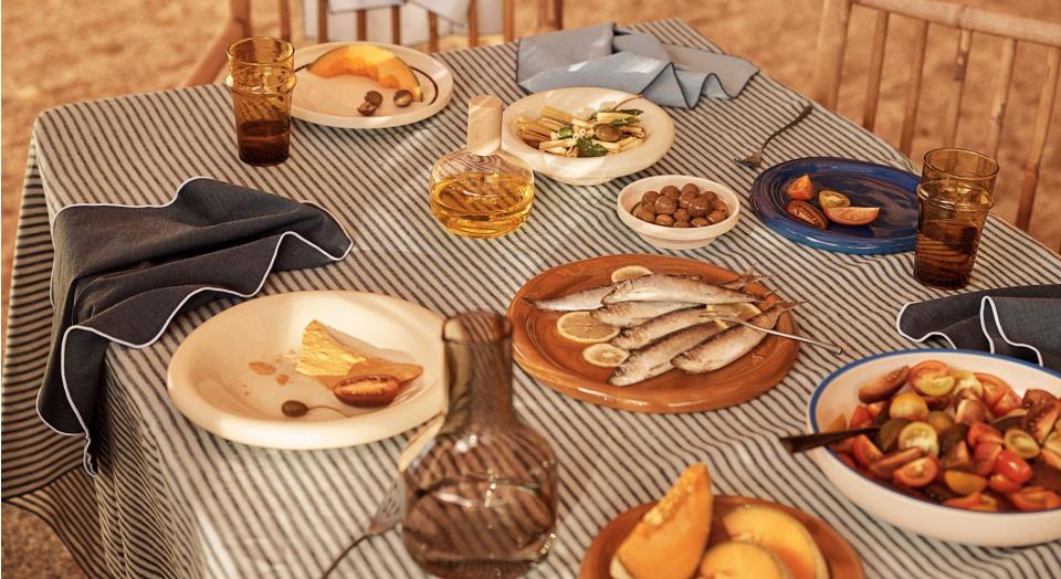 photo of styled table. ticking stripe linen table cloth with assorted coloured linen napkins, plates of fresh fruit, vegetables and fish sit on top