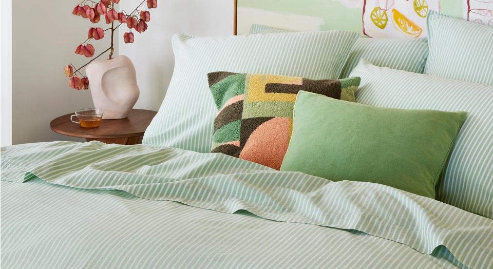 A close-up of a green striped bed. On top sits matching pillows and two accent cushions. Next to the bed is a wooden side table with a vase of dried flowers and a cup of tea.