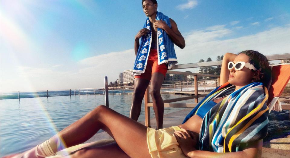 A young man and woman lounging by the beach with Sheridan beach towels draped over their shoulders