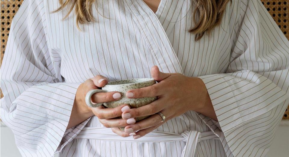 close up shot of blonde woman wearing striped cotton dressing gown, holding a cup of tea with her hands clasped around in. her nails are painted pink.