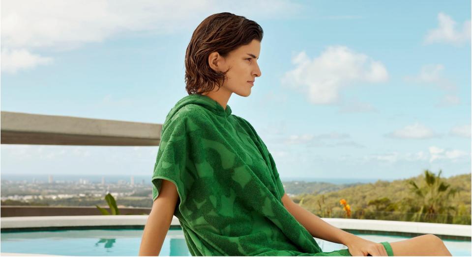 white woman with short brown hair sits in front of a pool, wearing a palm green hooded poncho with botanical design.