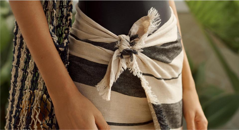 a close up shot of a white woman wearing a sarong as a skirt. it is tied on a knot at her waist. she also has a striped bag, slung over her shoulder resting against her hip. blurry green plants in the background.