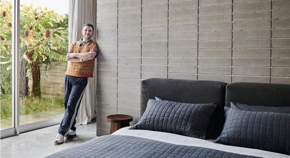 skincare specialist james vivian leans against exposed brick wall of his melbourne home, glass sliding door in background has view of green garden, foreground featured made up bed with quilted bed cover, pillowcases, silver sheet and velvet bedhead