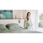 xanthe highfield, white woman with golden brown hair, stand next to her bed. she wears julip linen robe and is smiling, with hand on bed. bed is dressed in cactus abbotson linen.