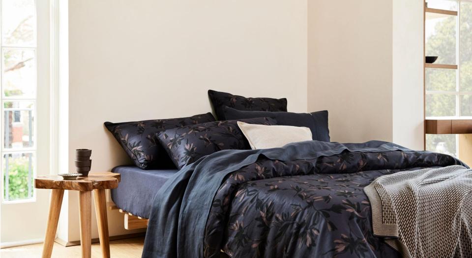 A bed dressed in black bed linen, sitting in a white bedroom beside a timber side table