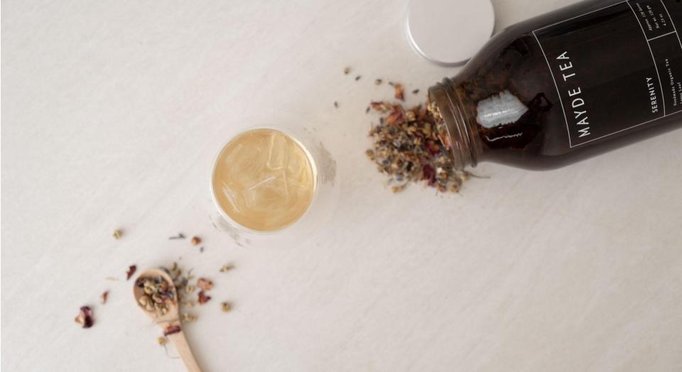 A cup of herbal tea sits next to a wooden spoon and glass jar. Tea leaves spill out from the jar and fill the spoon.