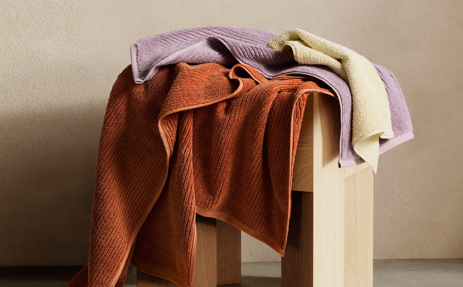 artful shot of light yellow, lilac and rust orange towels lying on wooden stool. neutral painted wall and marble floor.