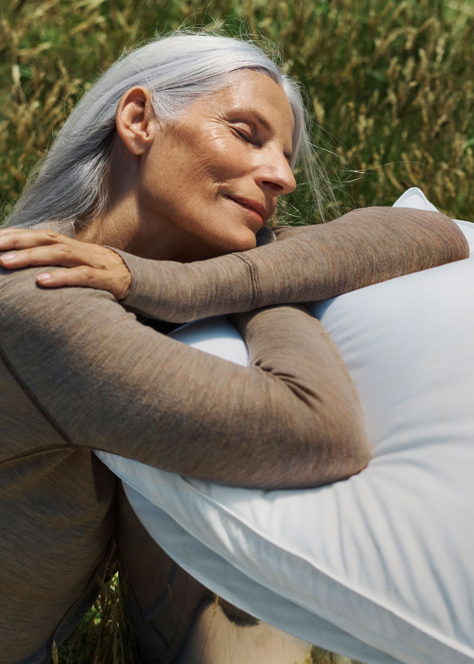 A woman with grey hair sitting in grass and leaning on a plush pillow.