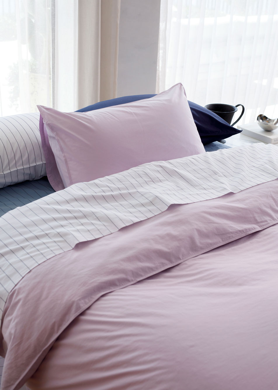 A bed dressed in Bayley Washed Percale bedding in the colours dusty mauve, white, steel blue and deep sea