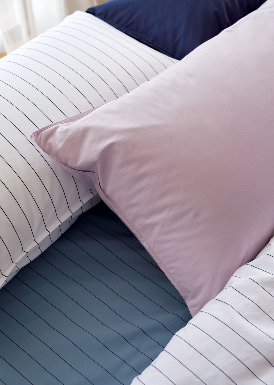 A close-up image of three pillows with Bayley Washed Percale pillowcases in dusty mauve, white stripe and deep sea.