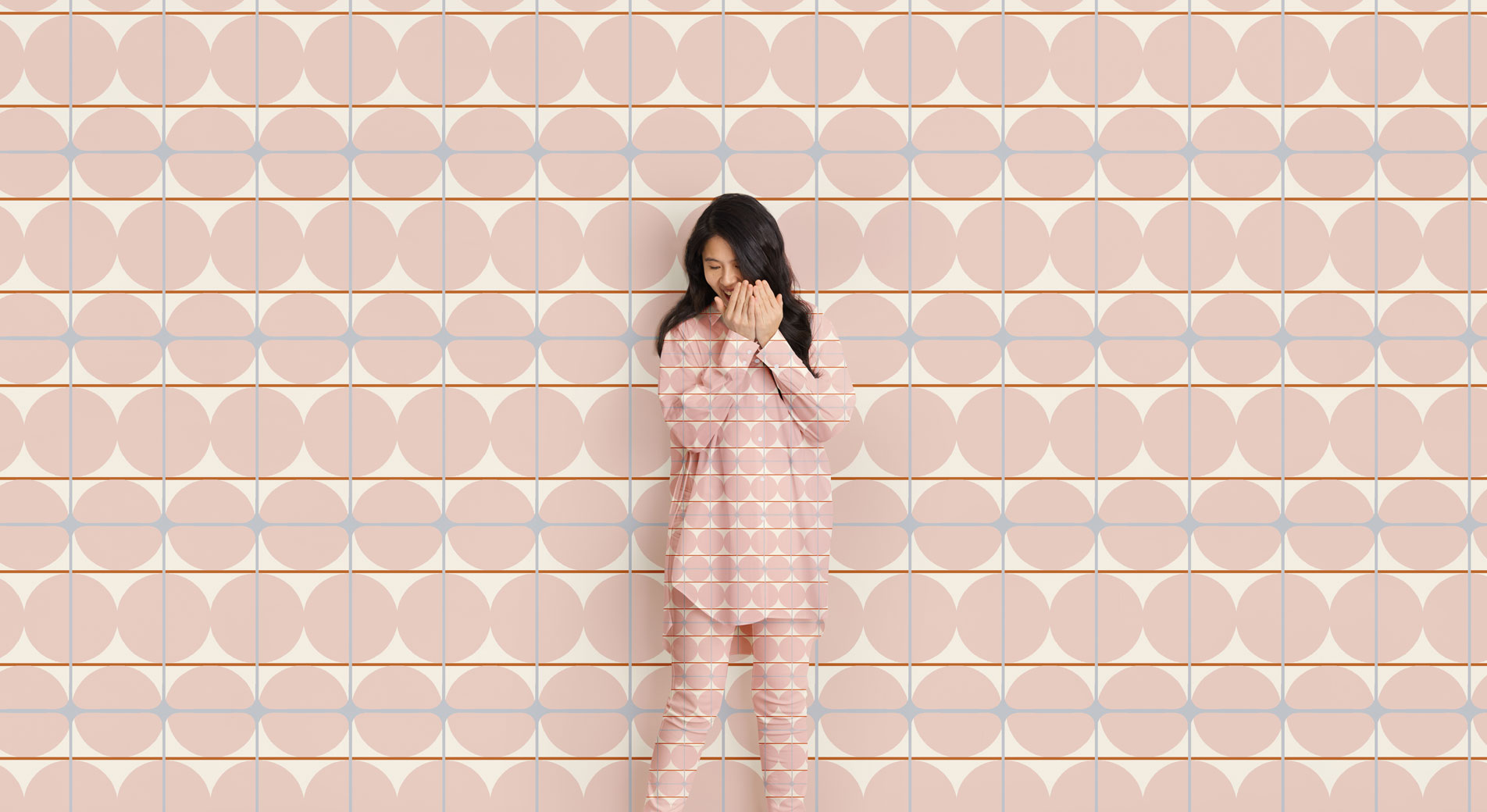 A woman stands, giggling and covering her face in front of a geometric print background. Her shirt and pants are in the same matching pattern.