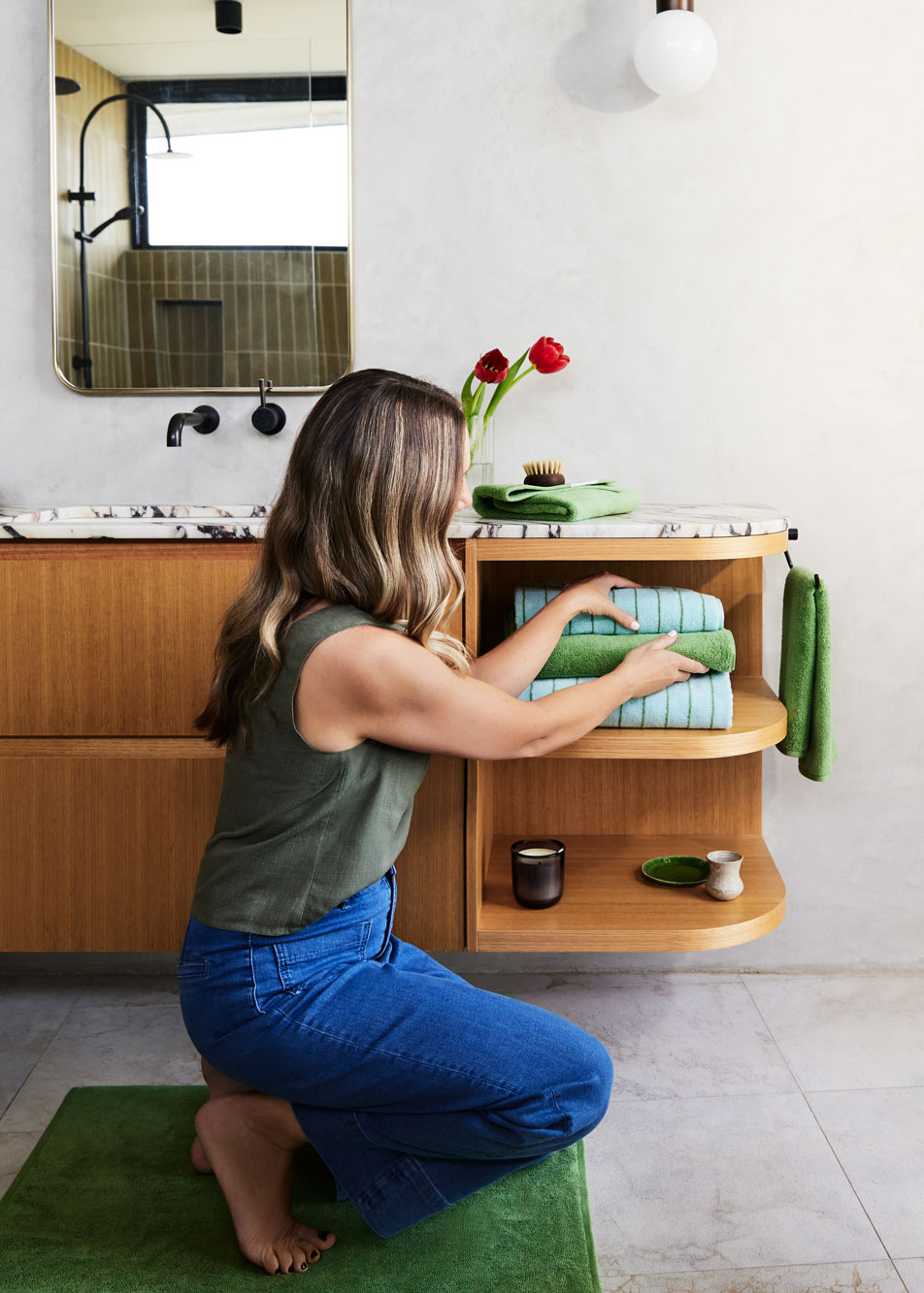 A woman crouching down in front of a modern bathroom vanity, tidying a pile of folded towels.