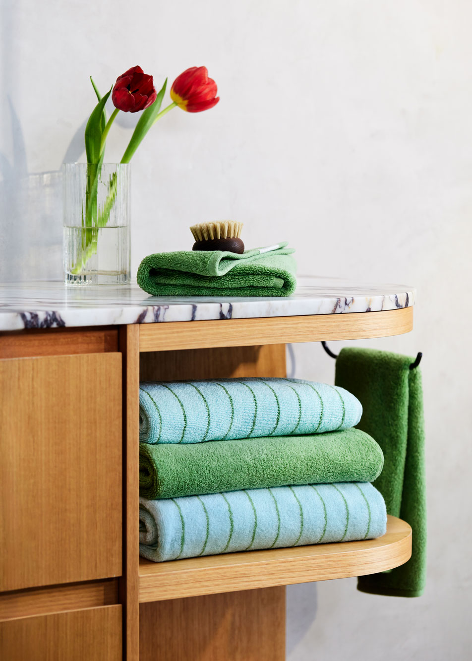 A timber and marble bathroom counter. On top is a glass vase holding two red tulips. Folded on top and in the shelves are Aven bath towels.