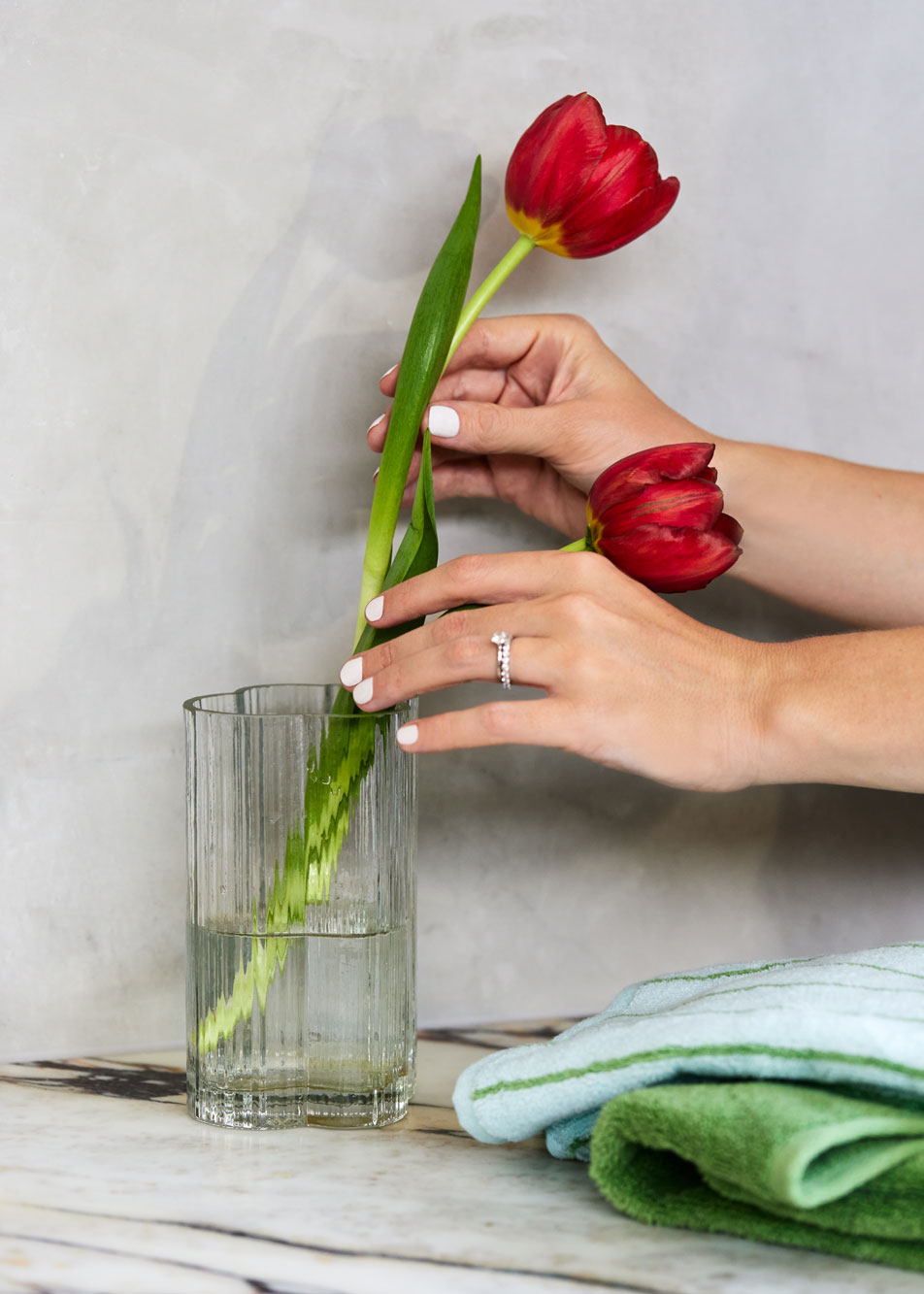 A pair of hands placing two red tulips in a glass vase. Beside the vase are two folded bath towels.