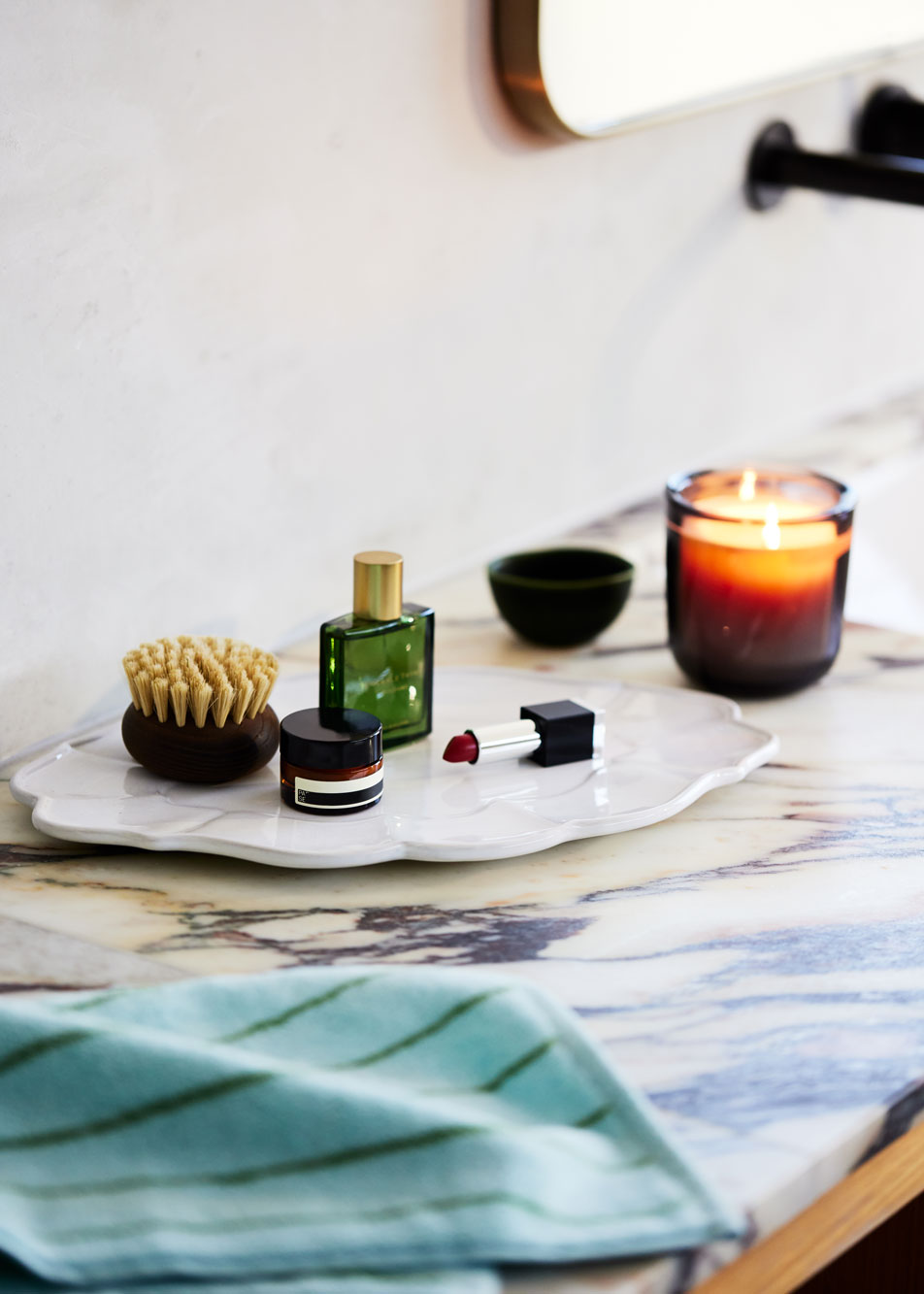 A marble benchtop. On top sits a ceramic tray with various makeup, skincare and perfumes. Beside it is a lit candle.
