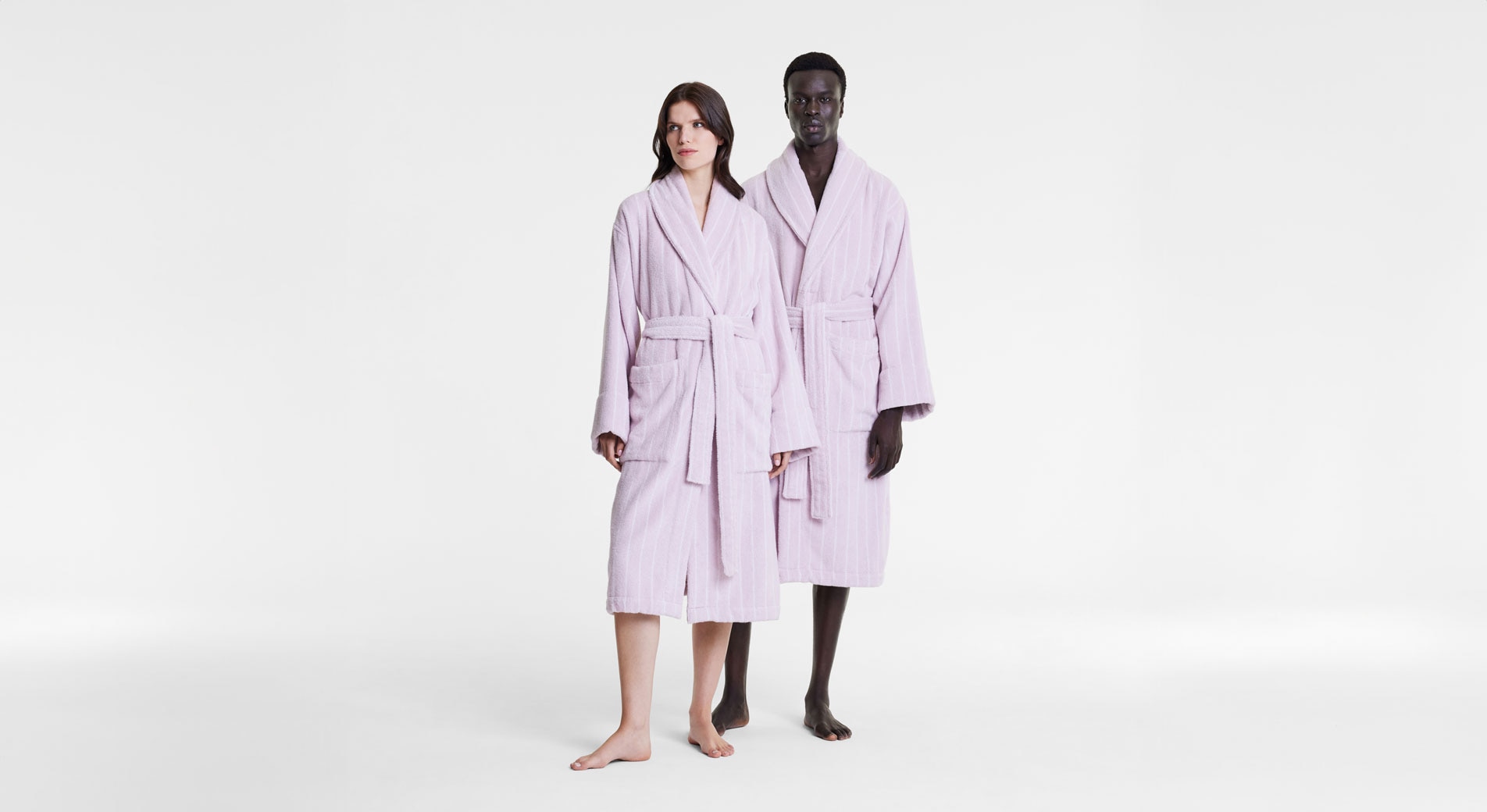 A man and woman stand side by side in front of a blank white set. Both are modelling Aven Stripe bathrobes in lilac.