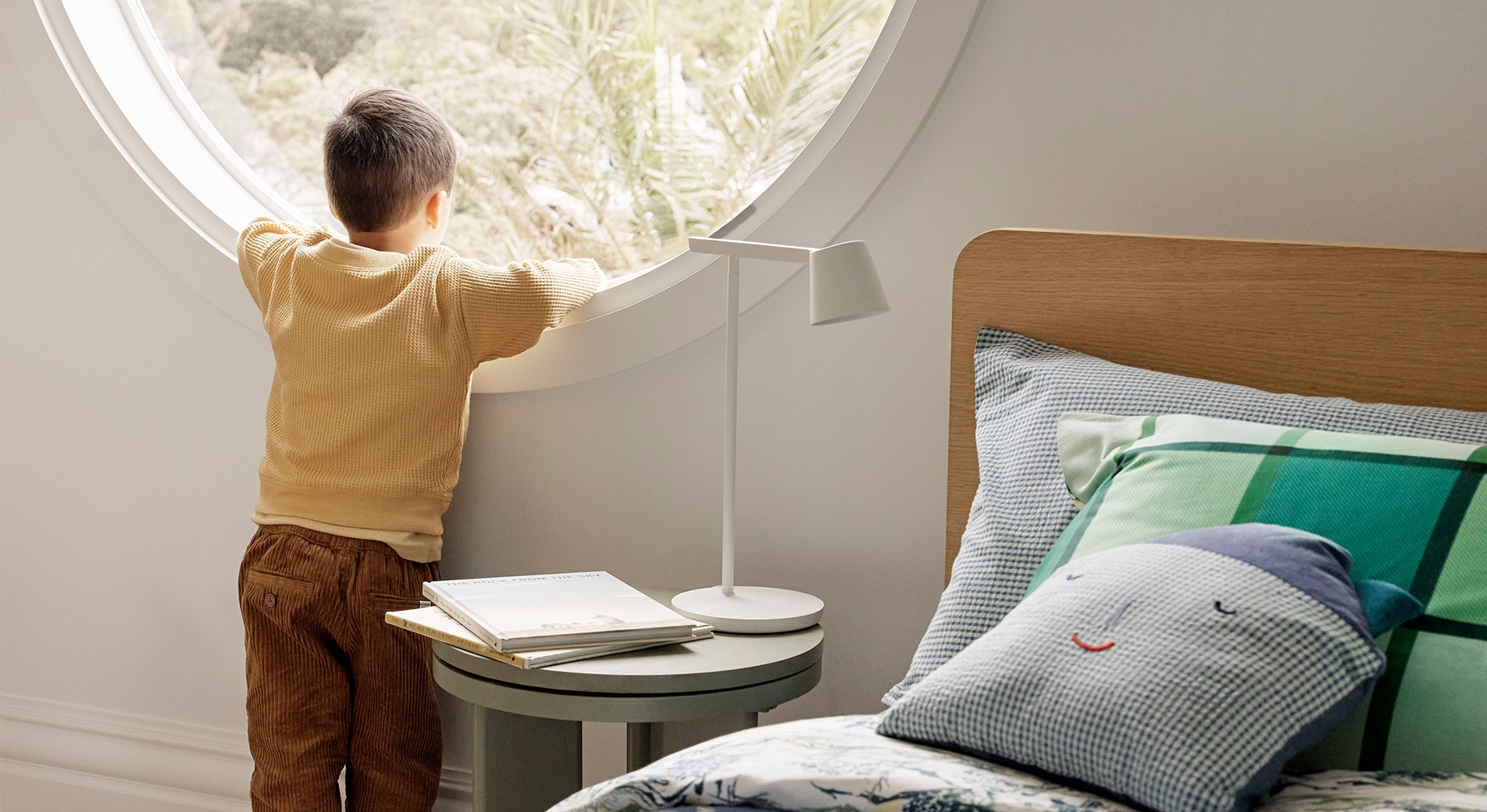 Moving your toddler into a big bed. A child stands in a bedroom looking out of a circular window at trees, next to them is a table with a lamp and books, as well as a bed with green and blue printed sheets and a house-shaped cushion