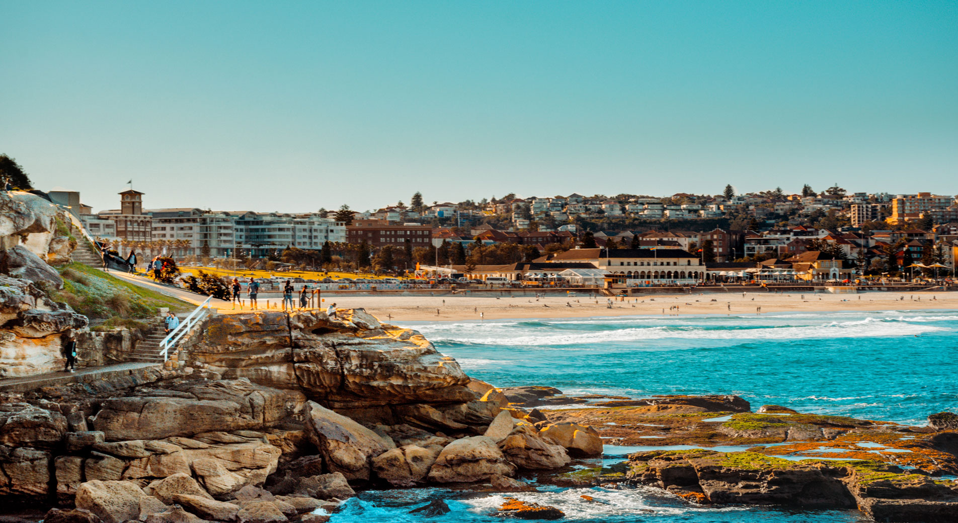 shout from rocky outcrop of bondi walk, looking back at bondi beach. the sun is setting off screen, throwing light on rocks, walkers and building, blue ocean waves crash on sandy shore.