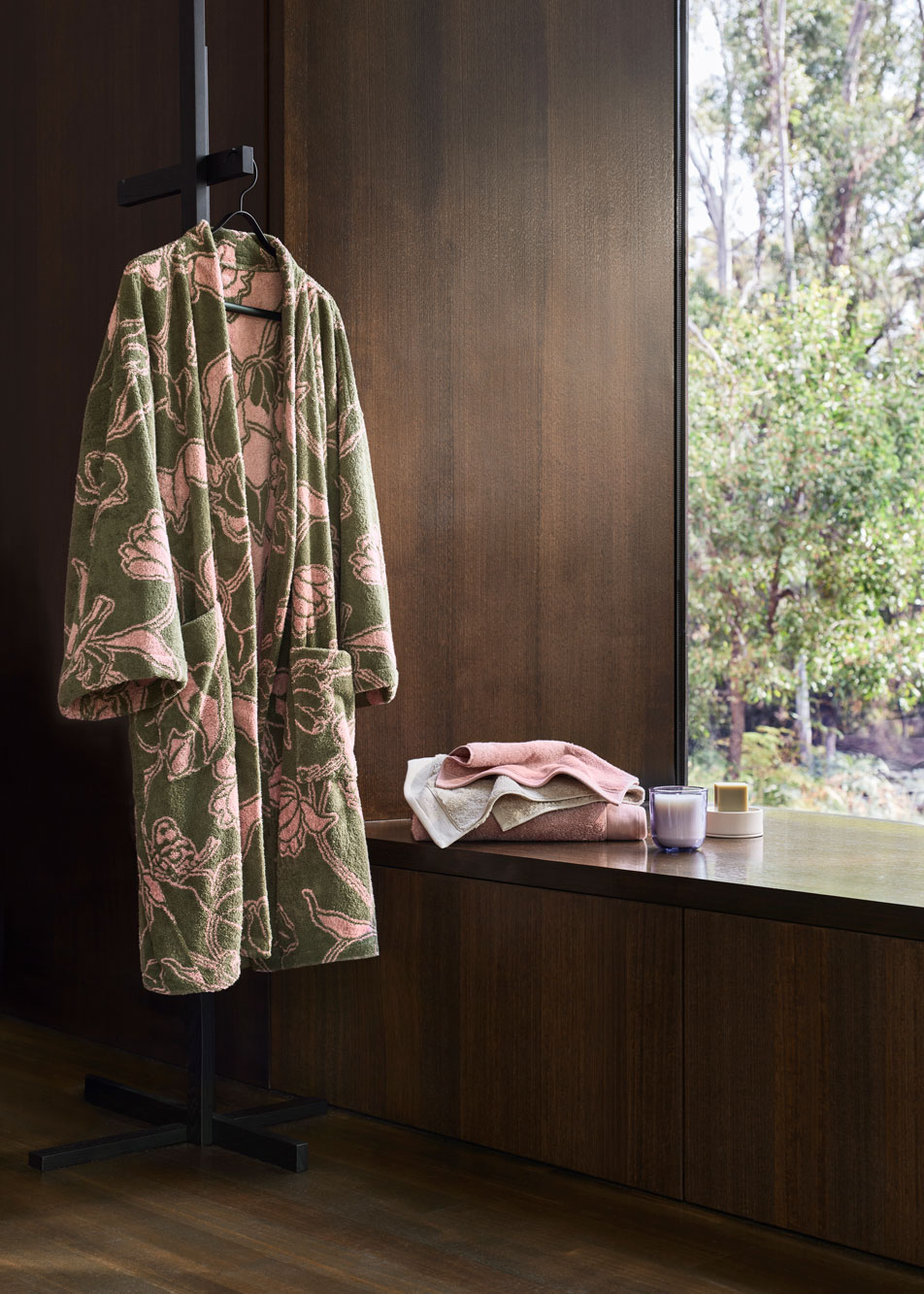 A modern, dark timber bathroom. A green and pink floral robe hangs from the wall, next to a stack of towels and a candle on the windowsill.