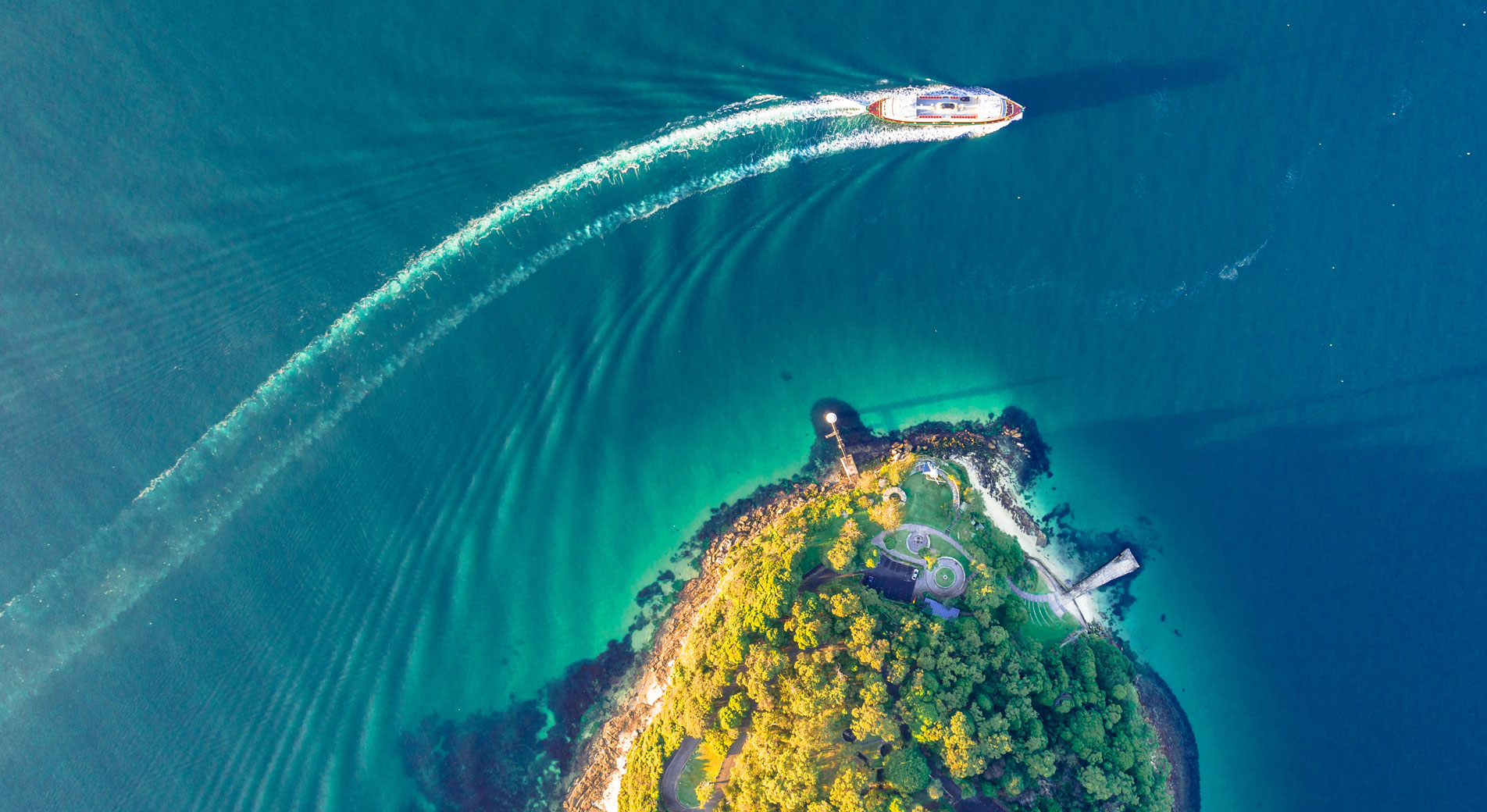 drone shot of bradley head ampitheatre. bush headland with small stretch of sand; bright blue sea with boat driving around it.