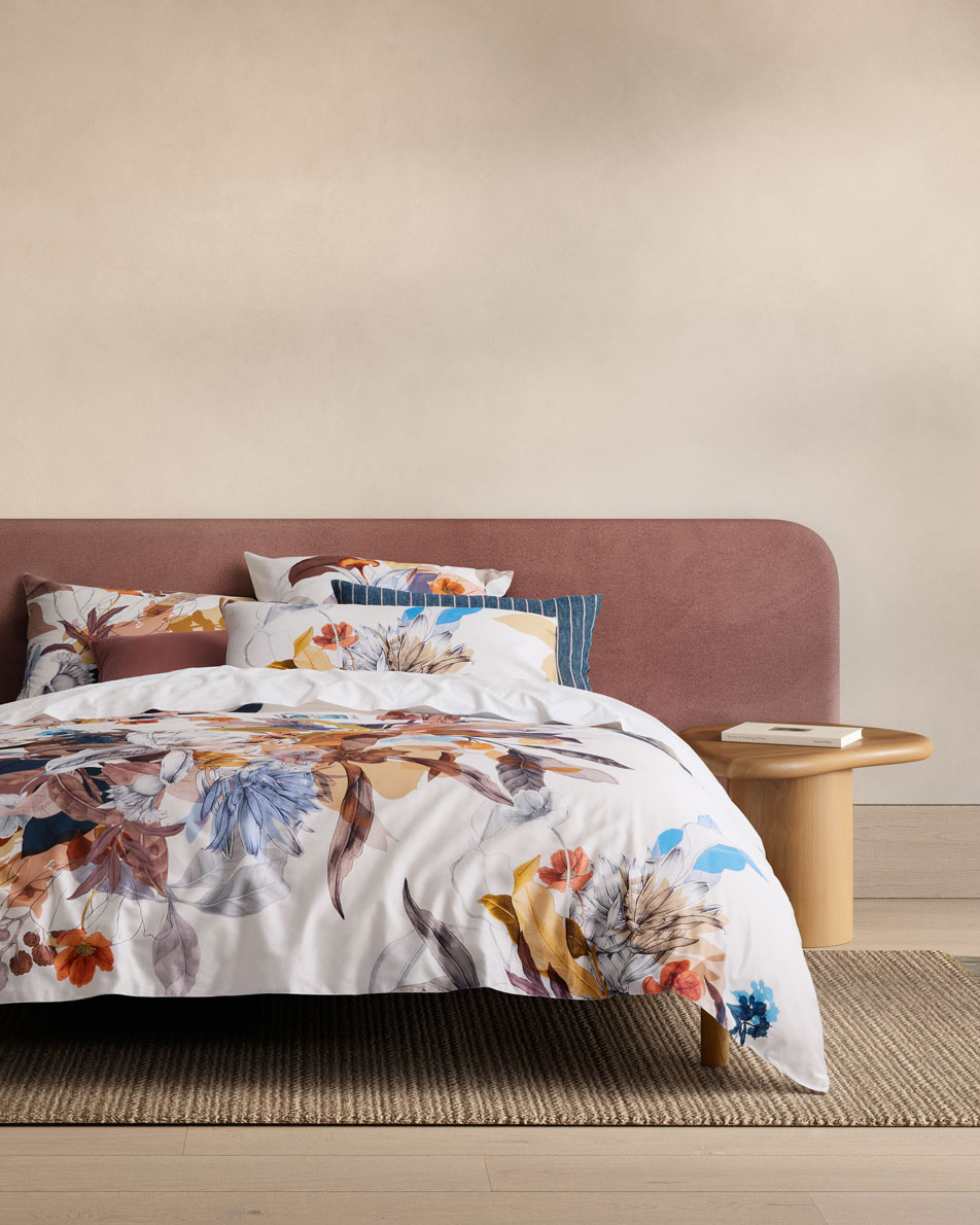 A colourful floral bed sits in a beige bedroom. Next to the bed is a timber side table.